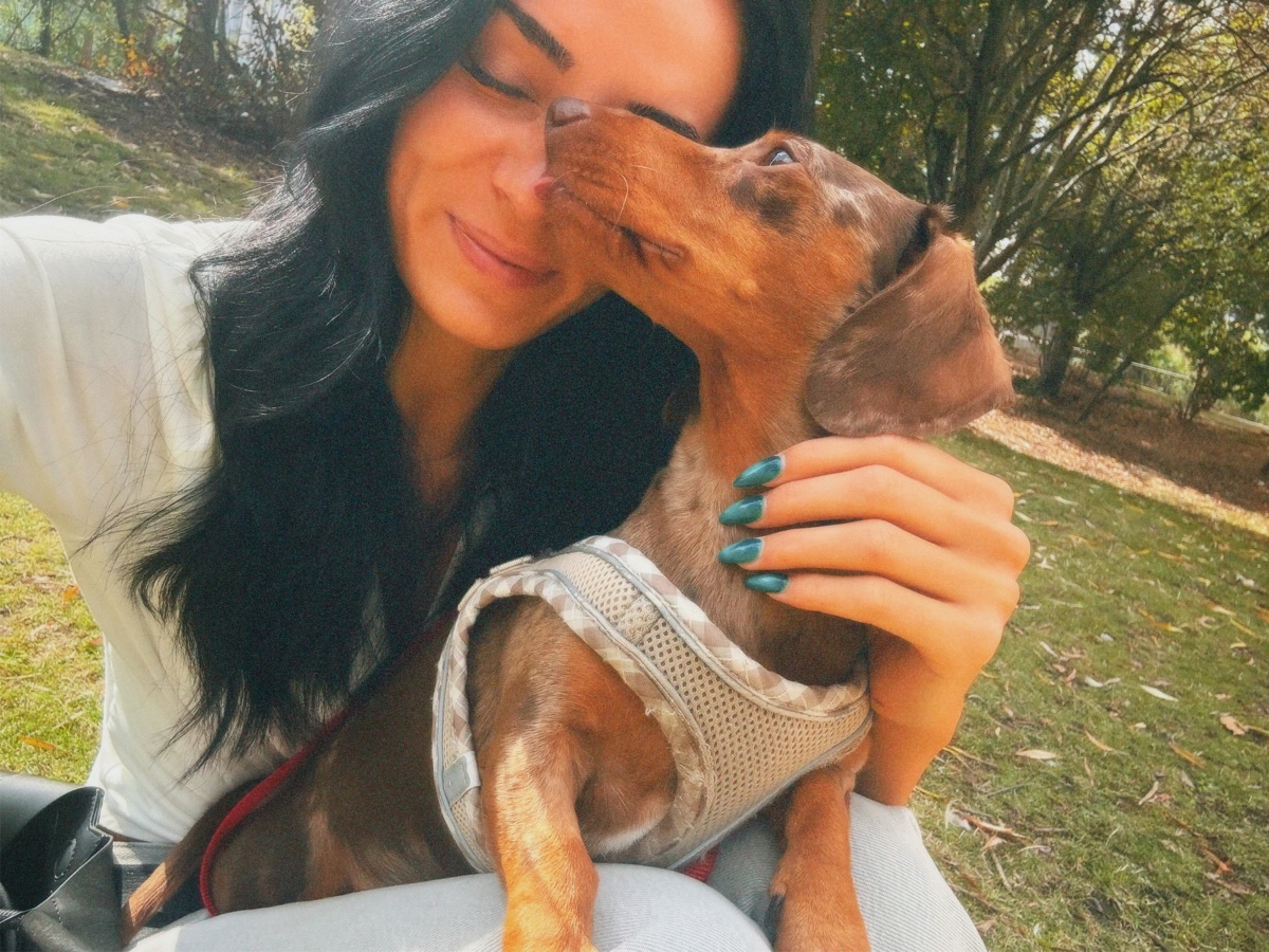 Lucy smiling with her eyes closed as a little dachshund in a harness kisses her cheek in an autumn park